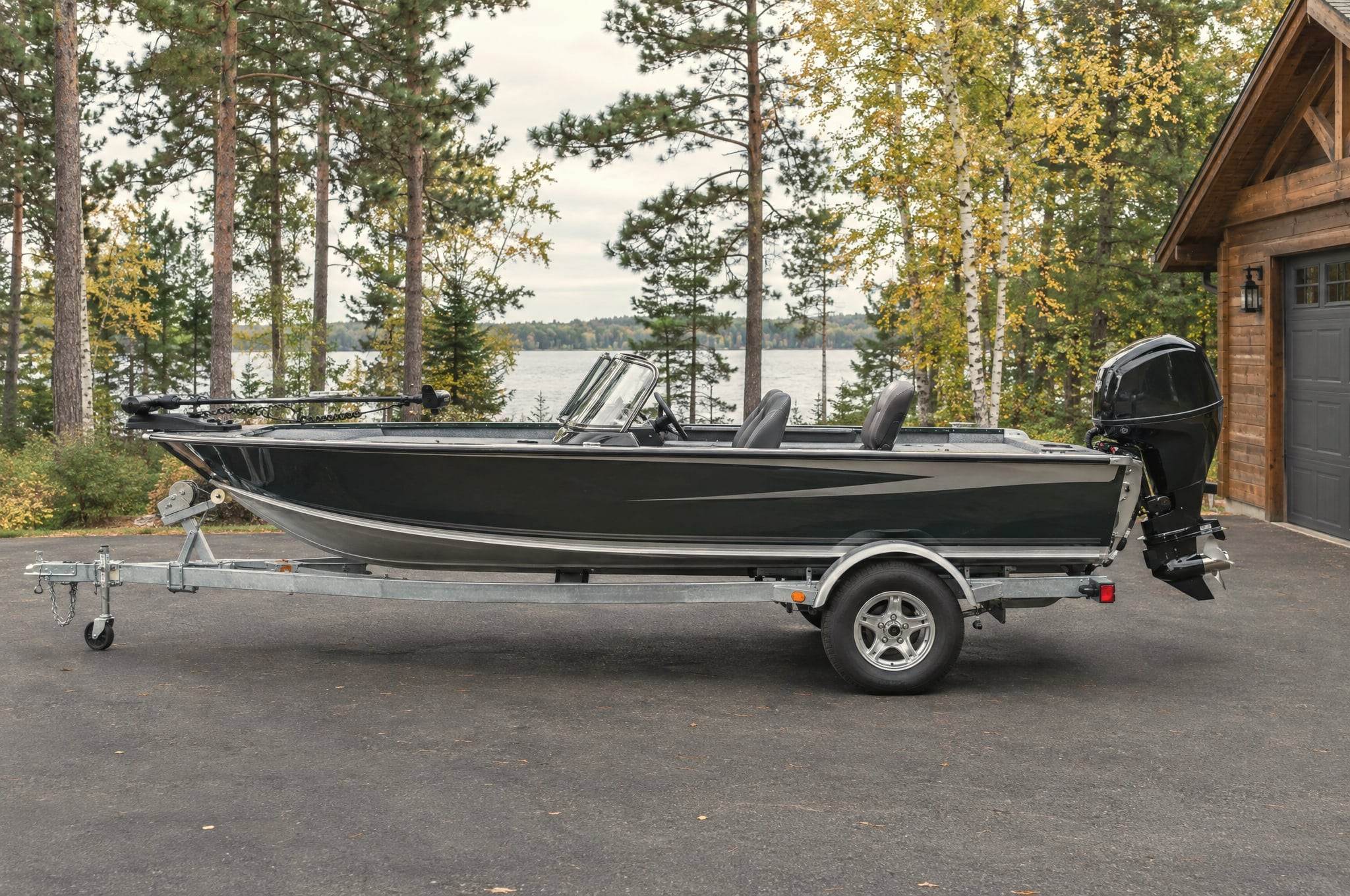 A fishing boat on a trailer in the drive beside a timber garage, with the lake through the pines